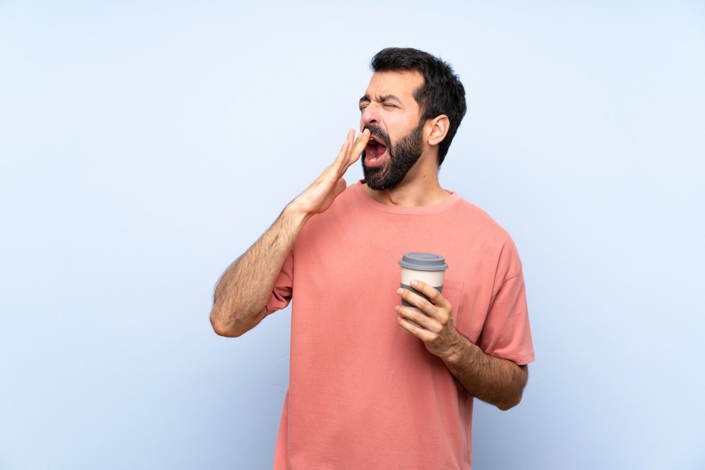 Young man with beard holding a take away coffee over isolated blue background yawning and covering wide open mouth with hand.