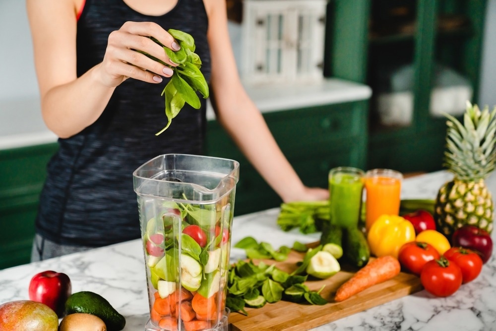 Young girl making green detox cocktail of fresh vegetables and fruits using blender in the kitchen.