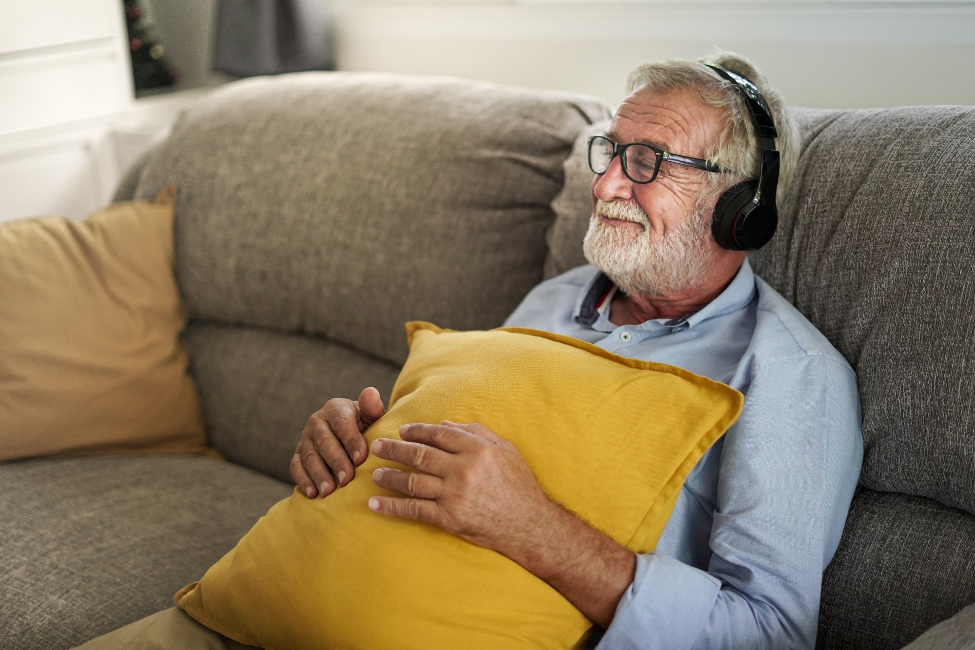 70-year-old man listening to music wearing headphones, hugging pillow, smiling